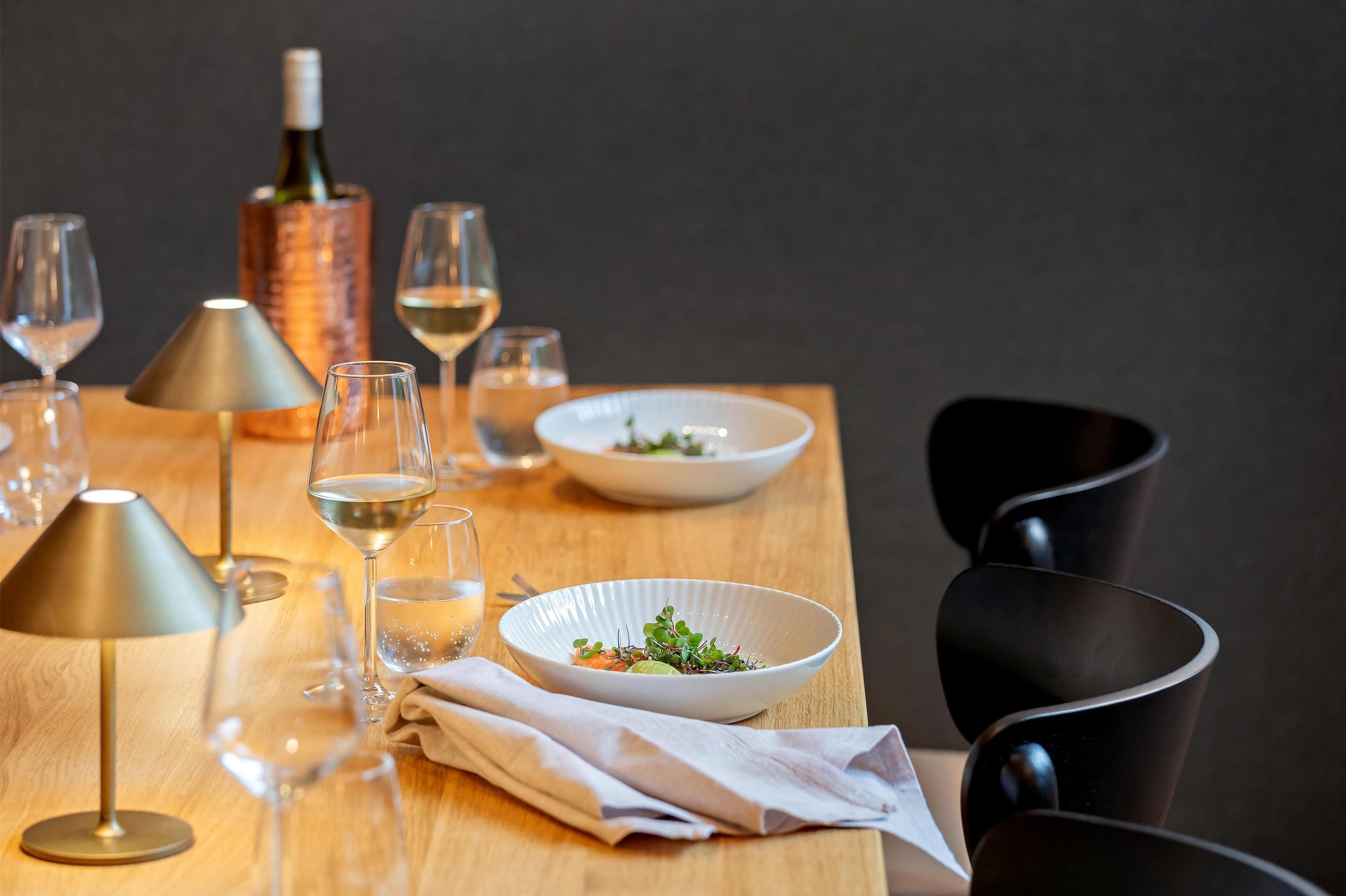 A dining table with a bottle of wine, glasses, a bowl of salad, and a lamp.