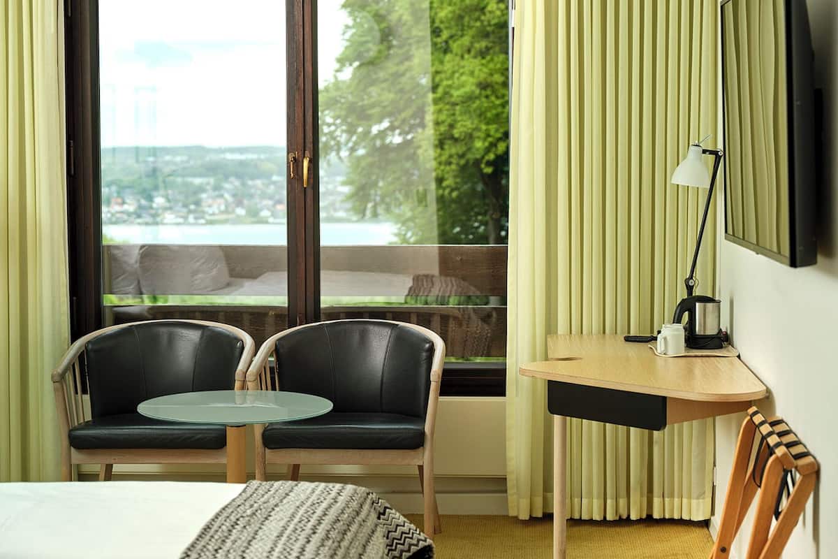 Ein Hotelzimmer mit Bett, zwei Stühlen, einem kleinen Tisch, einer Lampe und Blick auf eine Stadtlandschaft durch ein großes Fenster.