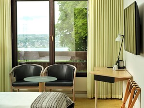 A hotel room with a bed, two chairs, a small table, a lamp, and a view of a cityscape through a large window.