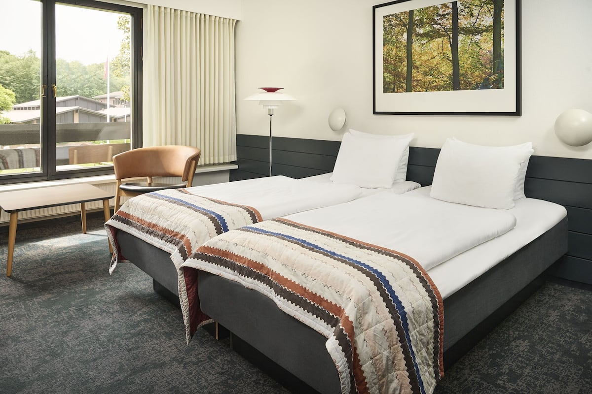 Ein Hotelzimmer mit einem großen Bett, einem Sessel, einem Schreibtisch, einem Fenster mit Vorhängen, einer Stehlampe und einem gerahmten Bild an der Wand.