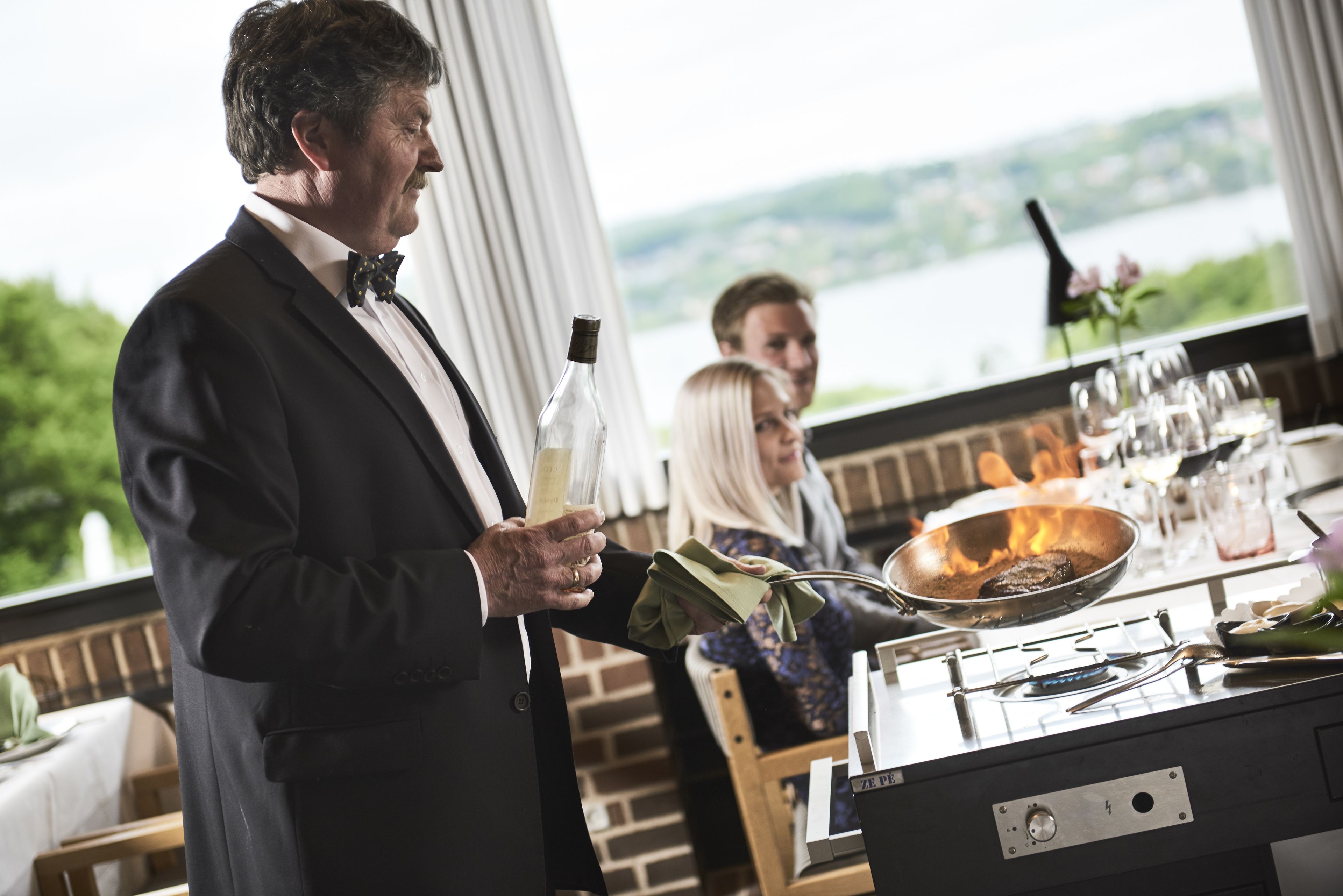 A person in a black suit holding a bottle and a napkin, standing near a stove with a pan on it.