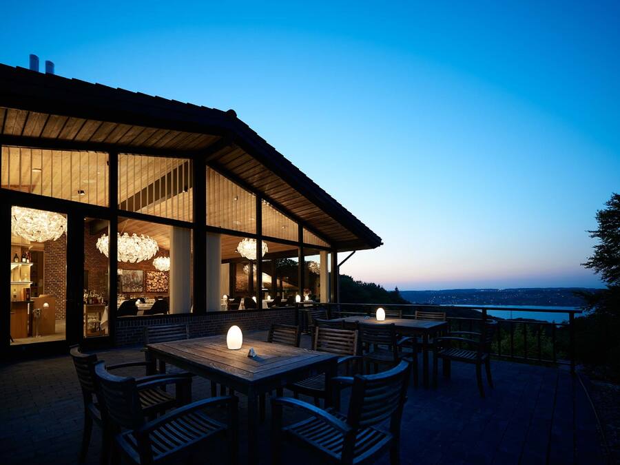 An outdoor dining area with wooden tables and chairs, large windows, and a view of the landscape.