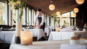 Un restaurante con mesas dispuestas para comer, un camarero atendiendo una mesa y grandes ventanales que dejan entrar luz natural.