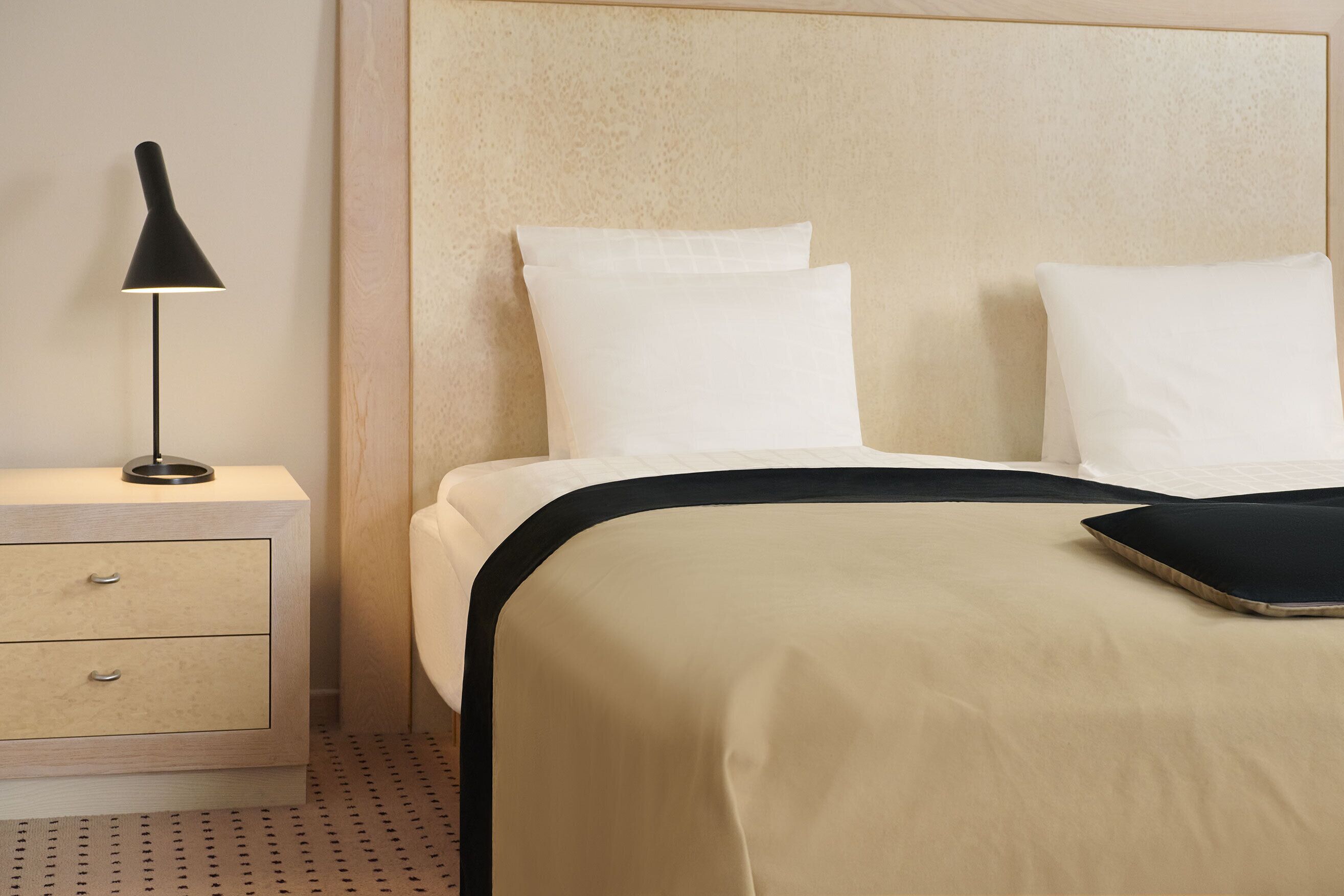 A neatly made bed with white linens and a black headboard, a bedside table with a lamp, and a patterned floor.