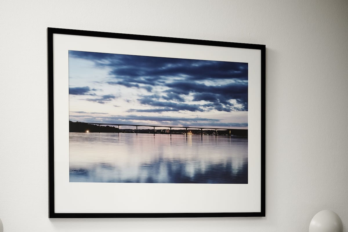 Ein ordentlich bezogenes Bett mit weißen Leinentextilien und einer Streifendecke, ein gerahmtes Foto einer Brücke über dem Wasser an der Wand und eine runde Wandleuchte.