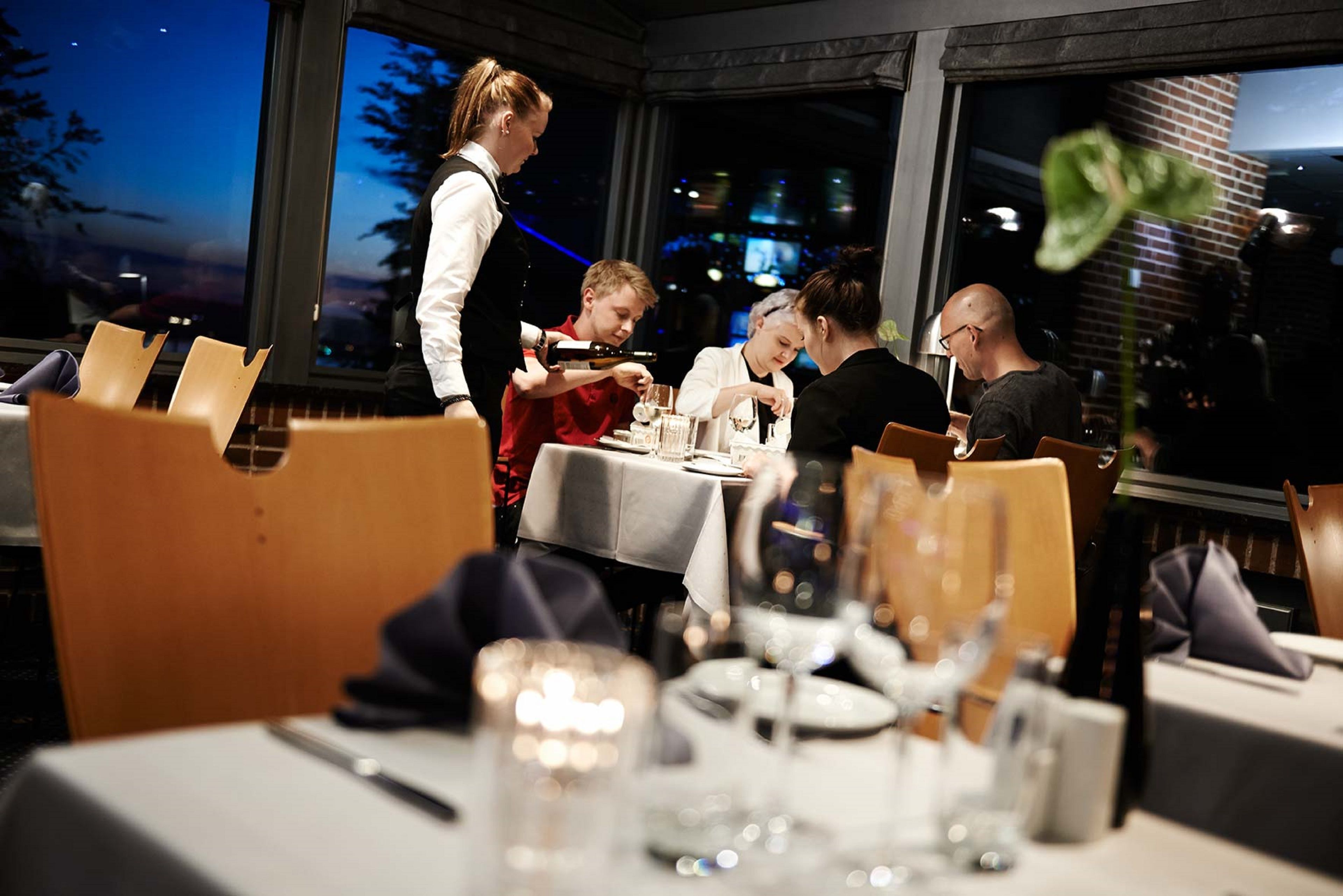 A restaurant with a table set for four people, a waiter attending to them, and a view of the cityscape through large windows.
