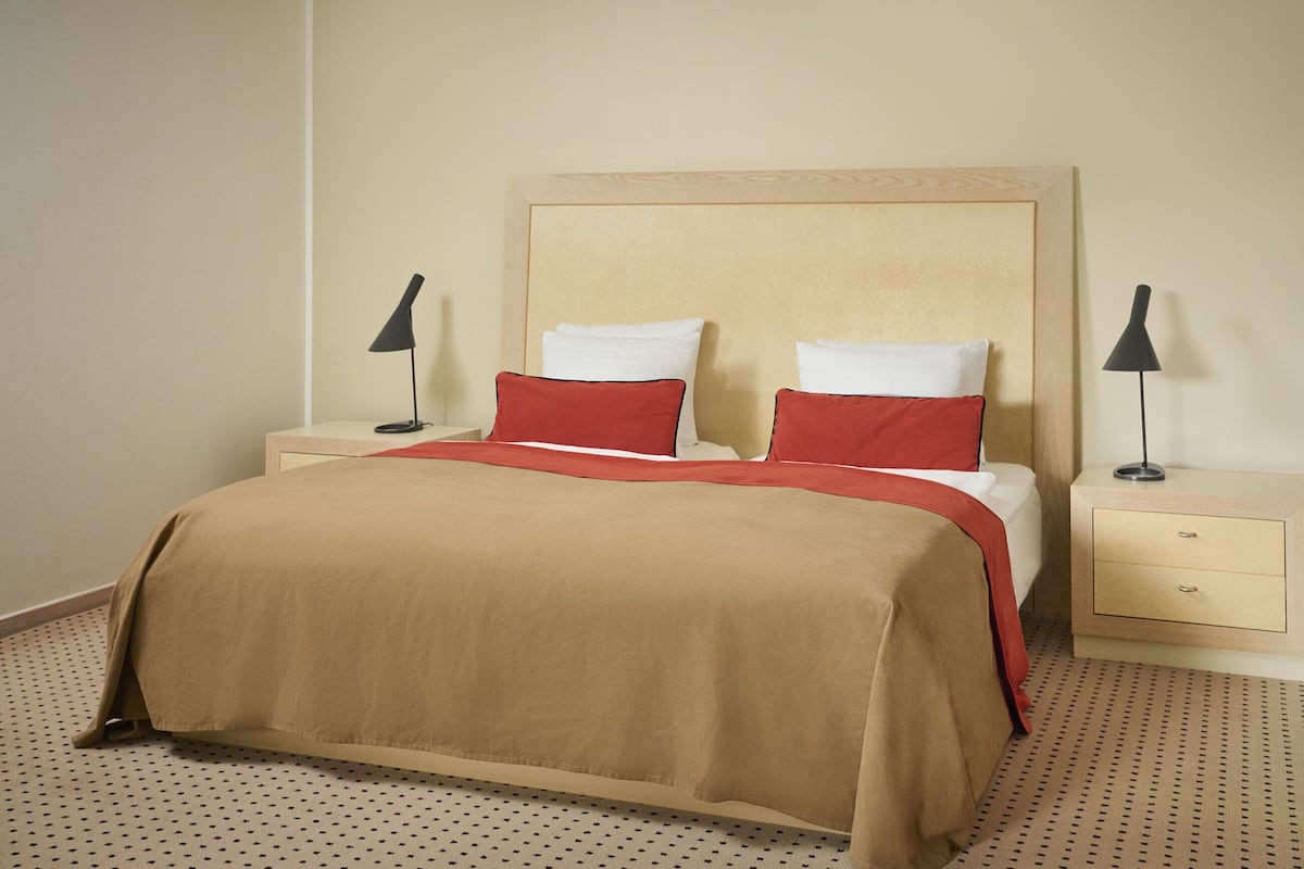 Ein ordentlich bezogenes Bett mit einer beige-roten Tagesdecke, zwei weiße Kissen und zwei rote Kissen. Zwei Nachttische mit schwarzen Lampen stehen daneben. Der Raum verfügt über einen beige farbenen Teppich mit Punktmuster.