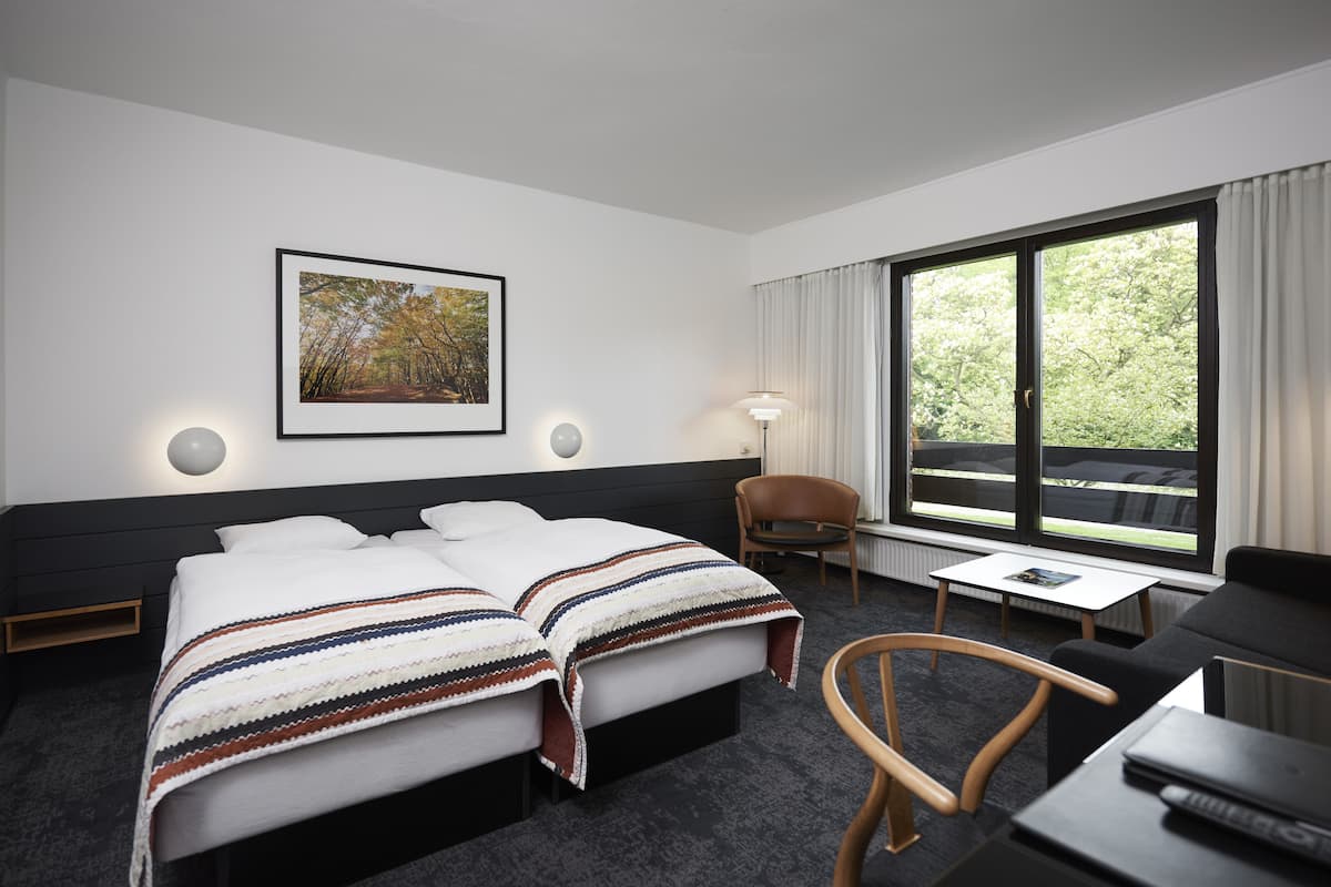 Ein Hotelzimmer mit zwei Betten, einem Schreibtisch, einem Stuhl, einem Fenster mit Vorhängen und einem gerahmten Bild an der Wand.