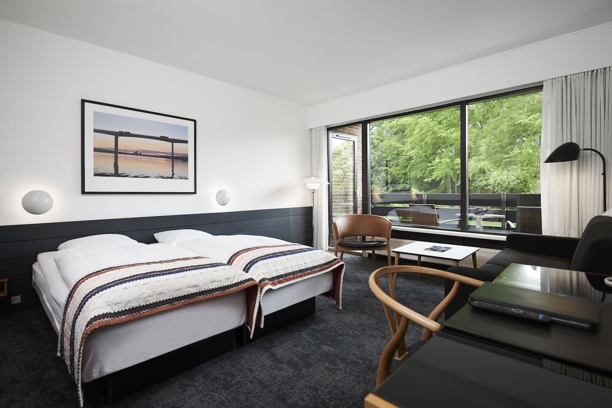Ein Hotelzimmer mit einem Bett, einem Schreibtisch mit einem Laptop, einem Stuhl und einem großen Fenster mit Blick auf Grünfläche.