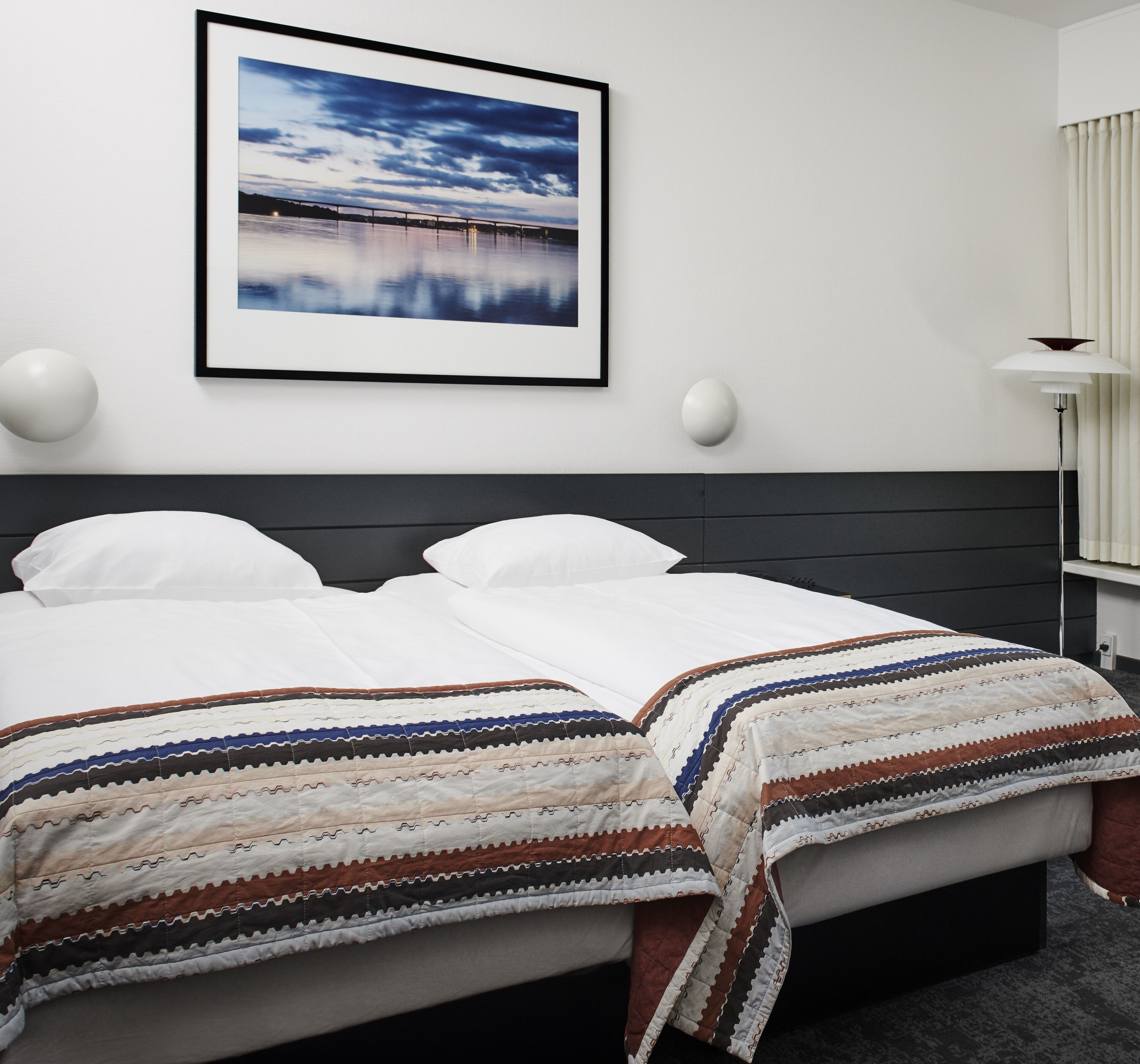 A neatly made bed with a striped blanket, a framed picture of a bridge on the wall, and a bedside lamp.