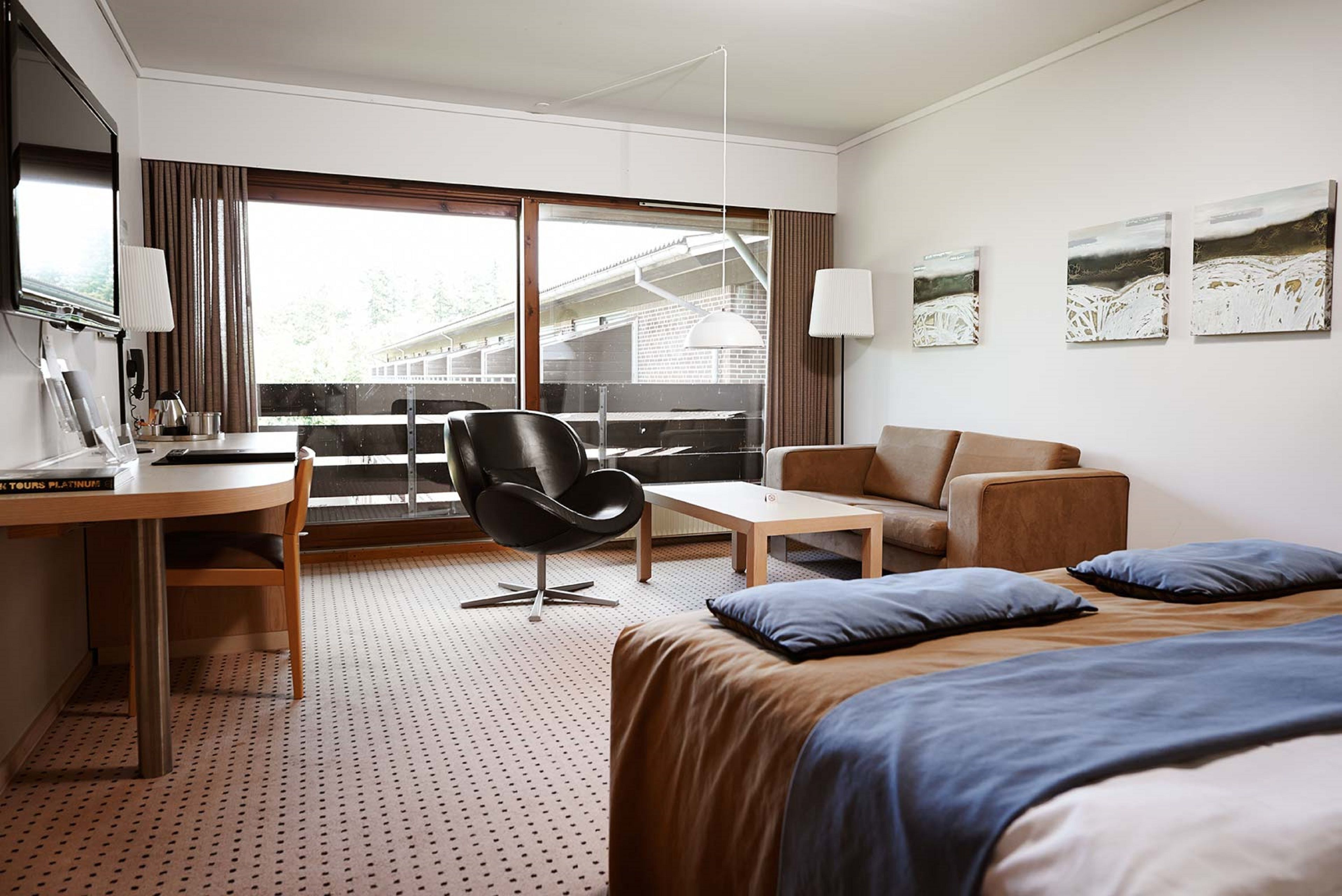 Habitación de hotel con cama, escritorio, silla, sofá y un amplio ventanal con vistas a edificios.