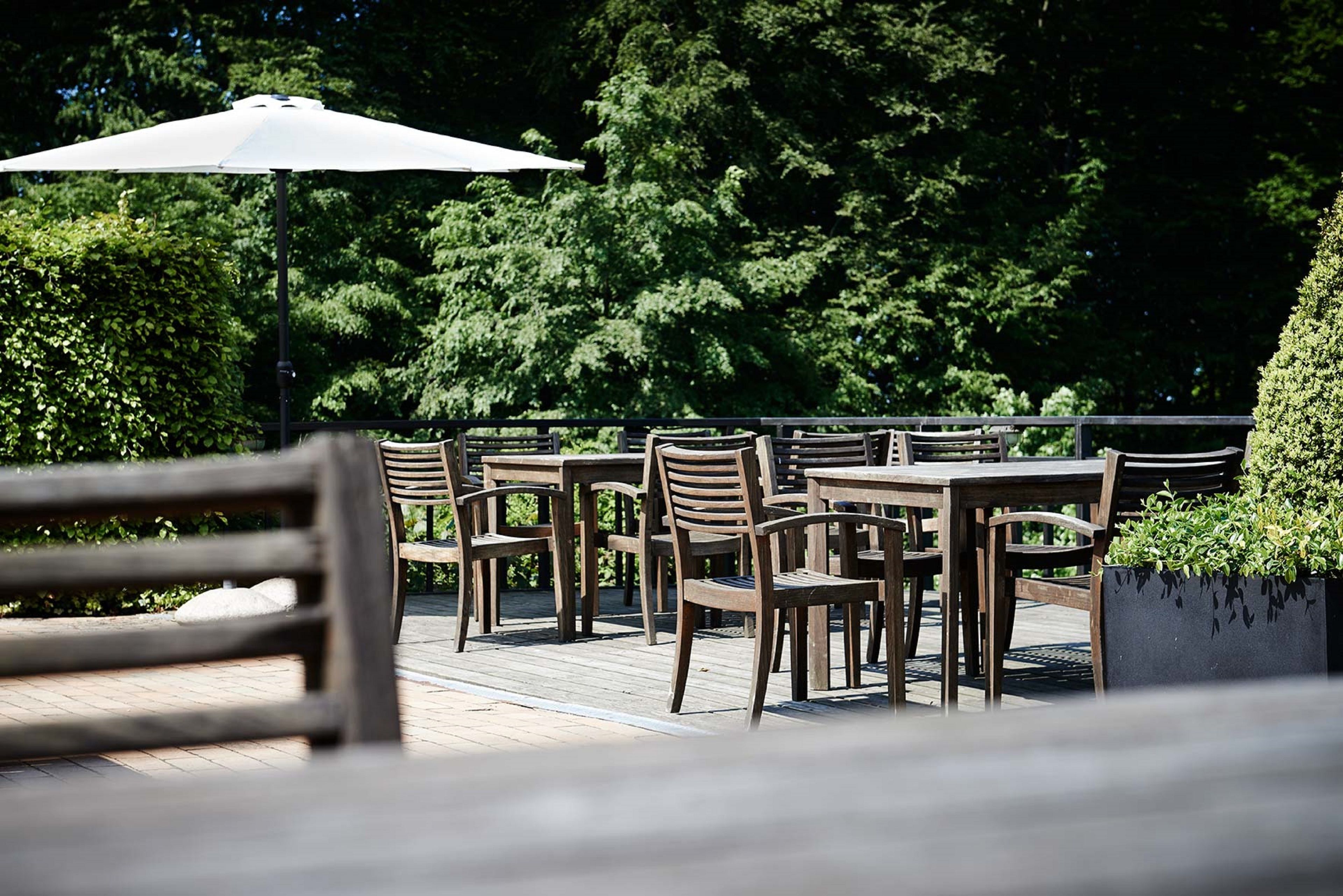 An outdoor patio with wooden tables and chairs, surrounded by lush greenery.