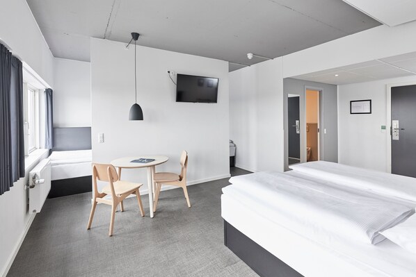 Quarto de hotel moderno com uma cama grande, uma mesa de trabalho com cadeira, uma TV fixada na parede e uma janela com cortinas.
