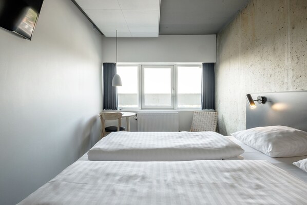 Habitación de hotel con cama, escritorio, silla, ventana con cortinas y lámpara fijada a la pared.