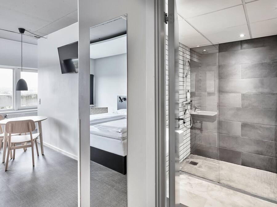 Habitación de hotel moderna con separador de vidrio, una cama, un escritorio con silla, zona de ducha con paredes de azulejos y vistas a la ciudad a través de una ventana.