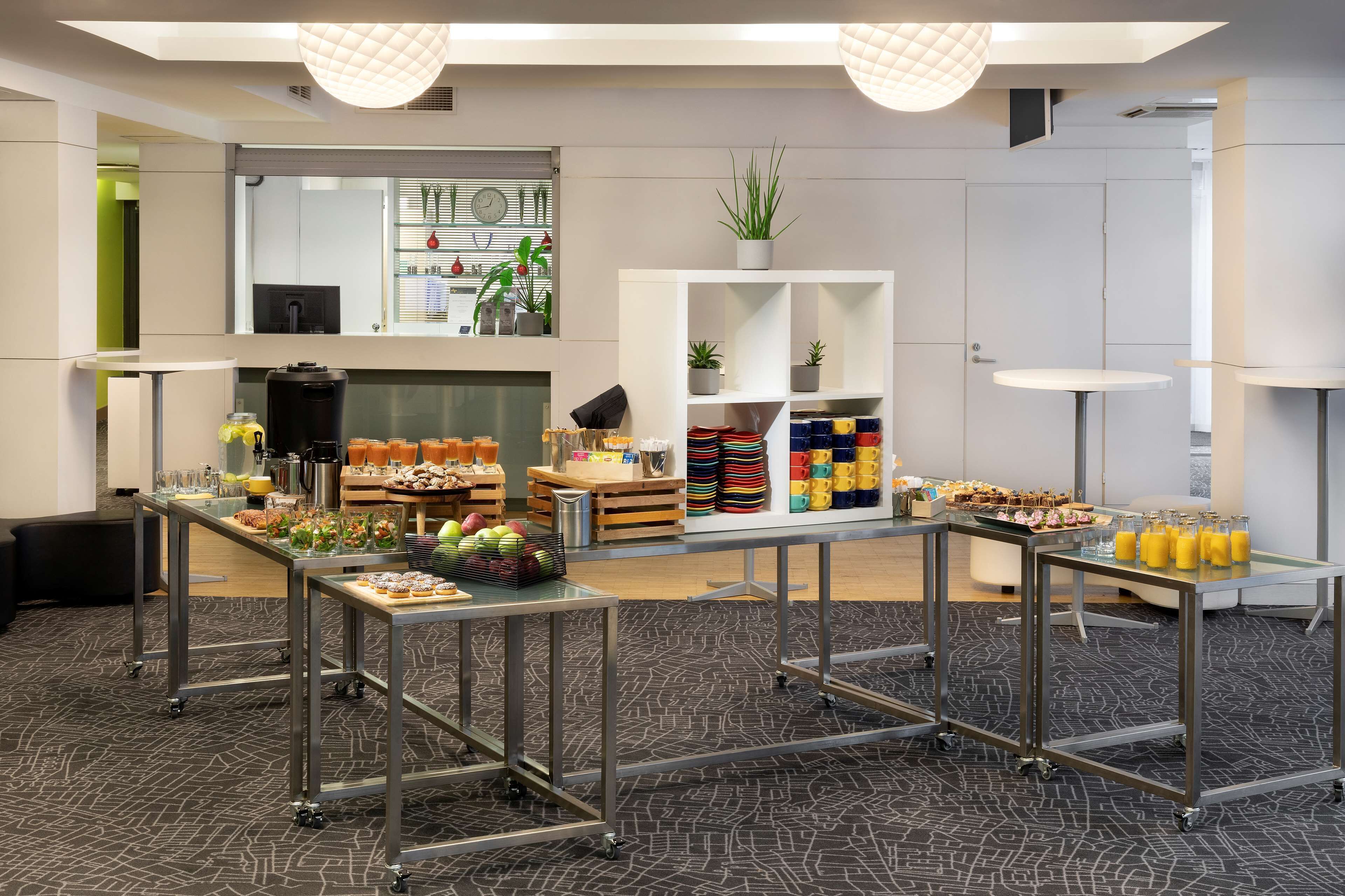 Habitación de hotel moderna con kitchenette, zona de comedor con comida tipo buffet y un amplio ventanal con persianas.
