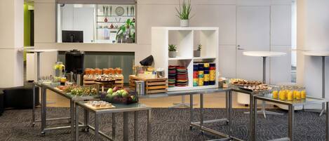 Habitación de hotel moderna con kitchenette, zona de comedor con comida tipo buffet y un amplio ventanal con persianas.