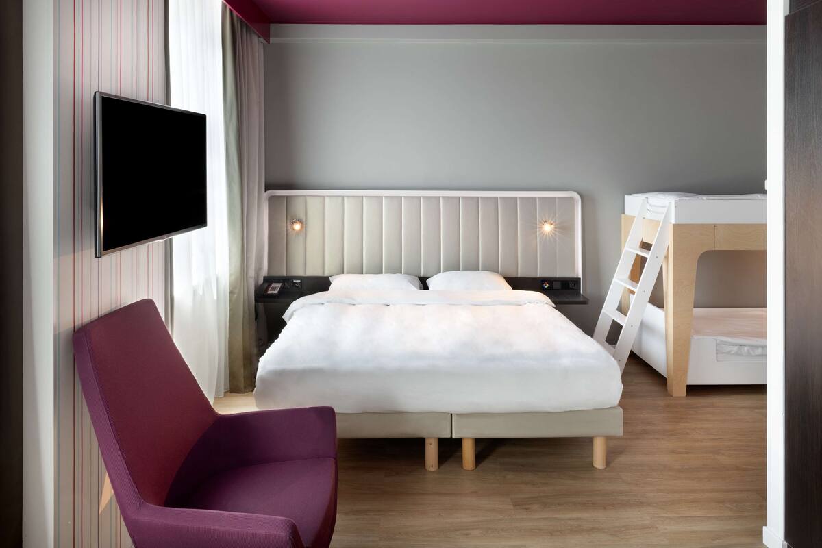 A modern hotel room with a bed, a TV mounted on the wall, a chair, and a small desk.