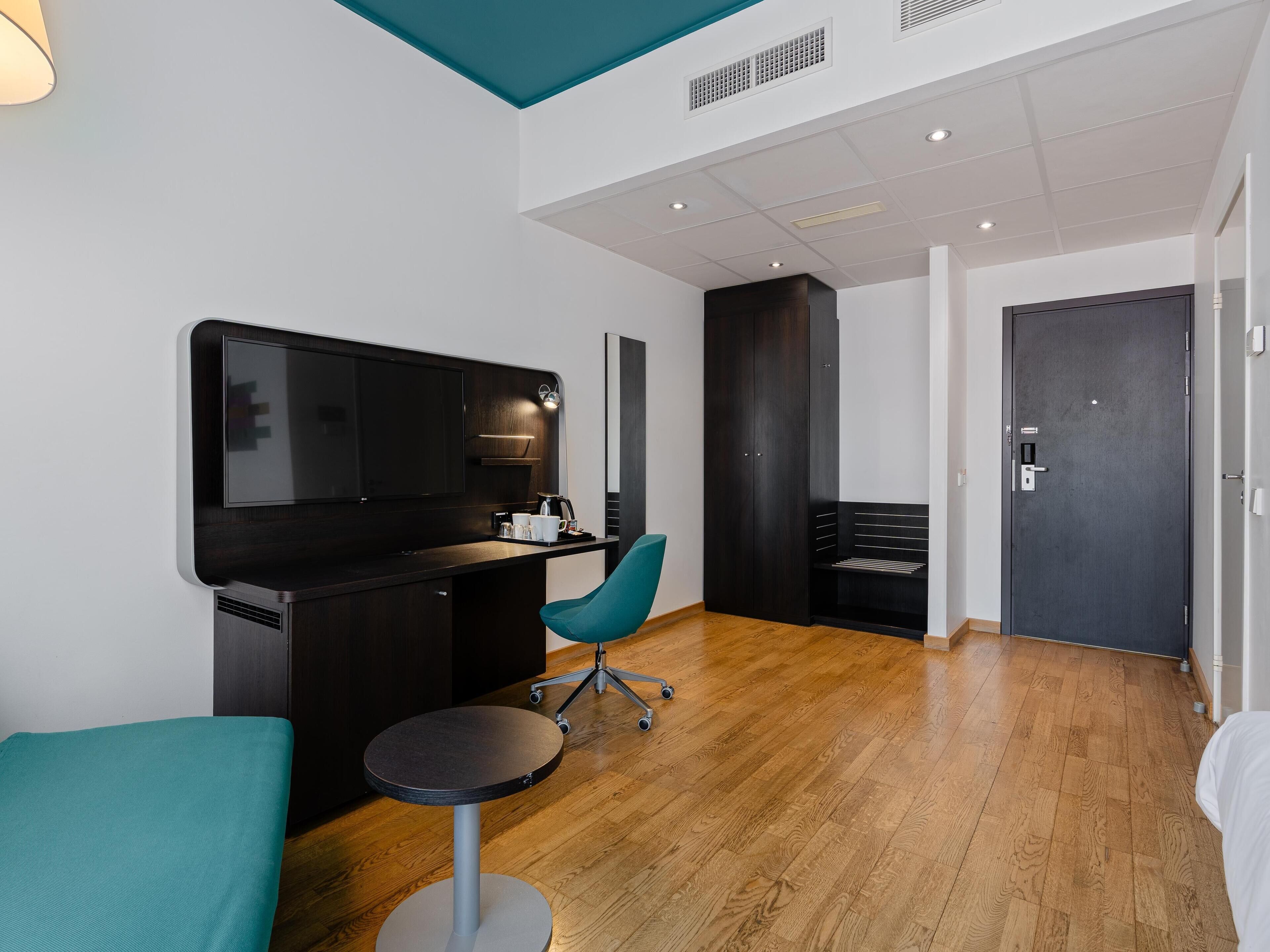 Une chambre d’hôtel moderne avec un bureau en bois foncé, une chaise turquoise, une table ronde, un grand téléviseur à écran plat et une armoire sombre.