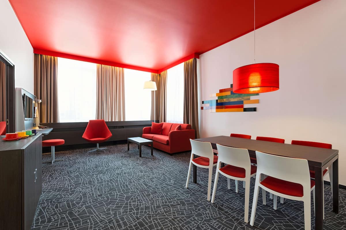 A modern hotel room with a red ceiling, a dining area with a long table and red chairs, a sofa, and a TV on a desk.