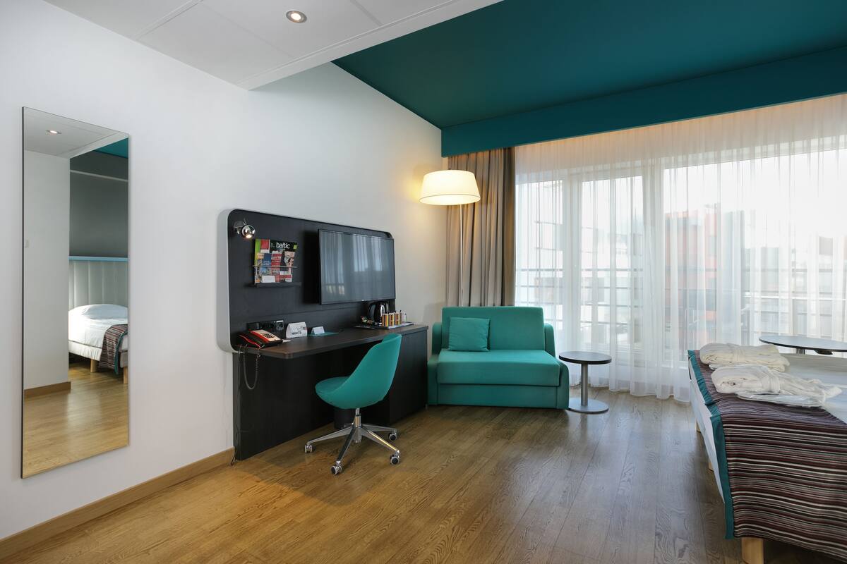 A hotel room with a wooden floor, a teal sofa, a desk with a chair, a bed with a striped blanket, and a large window with sheer curtains.