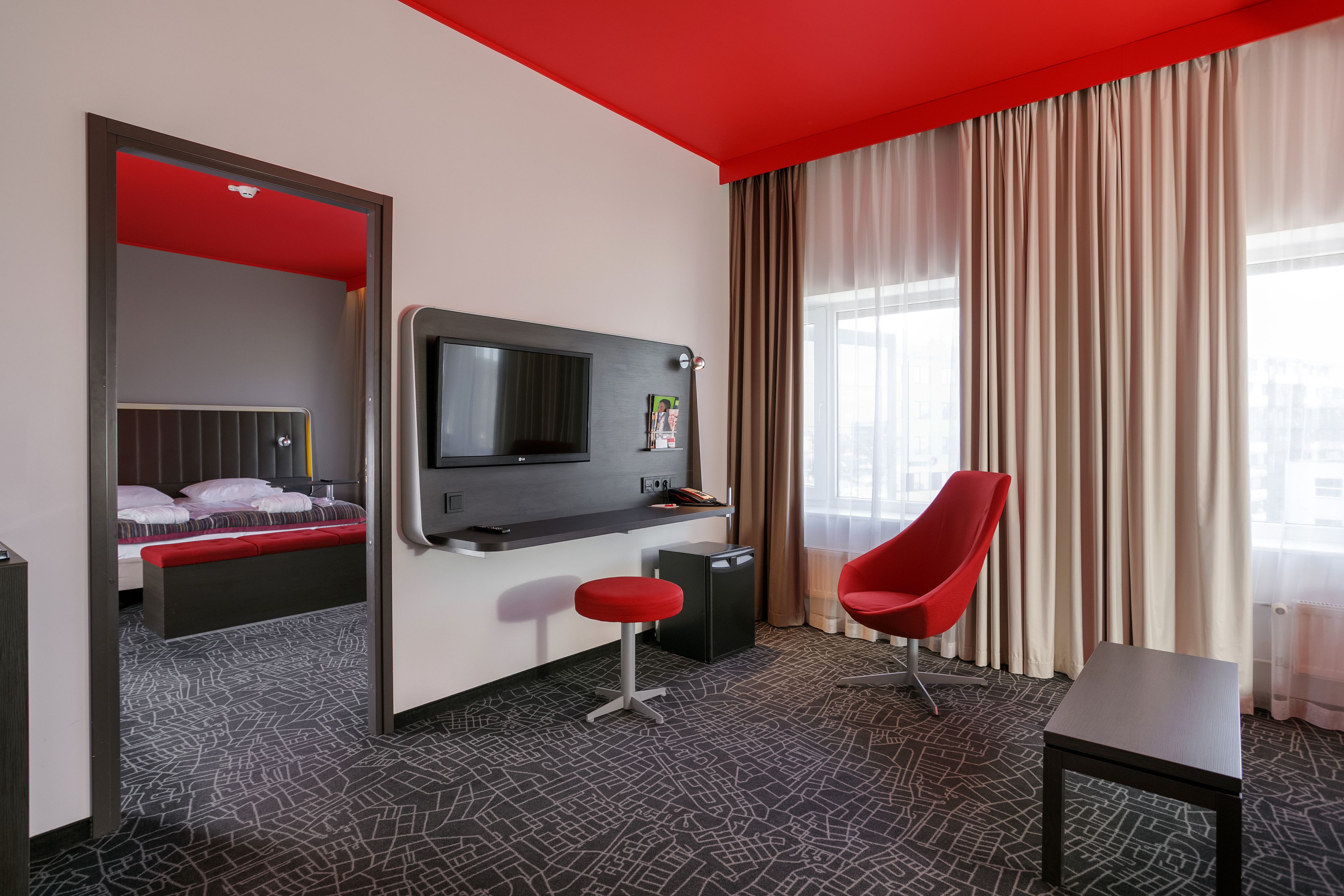 A hotel room with a bed, a desk with a TV, a red chair, and a small table.