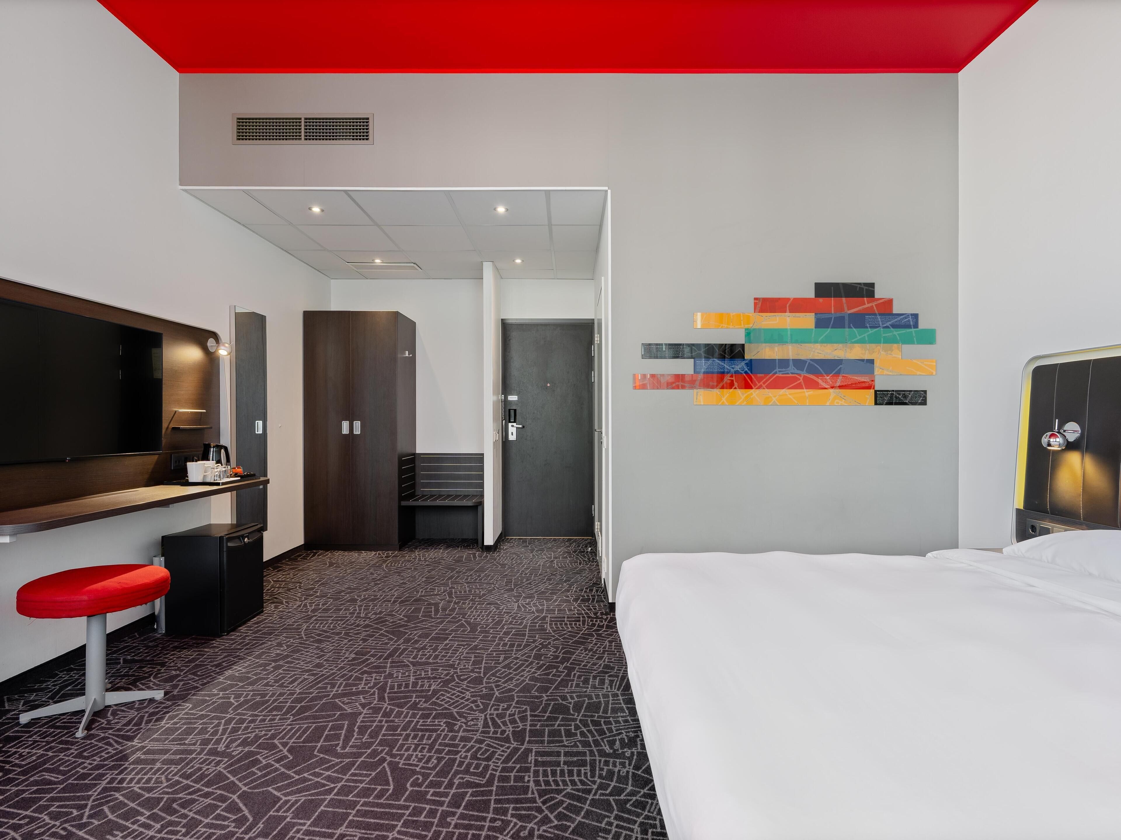A hotel room with a bed, a desk with a chair, a TV, a wardrobe, and a colorful abstract art piece on the wall.
