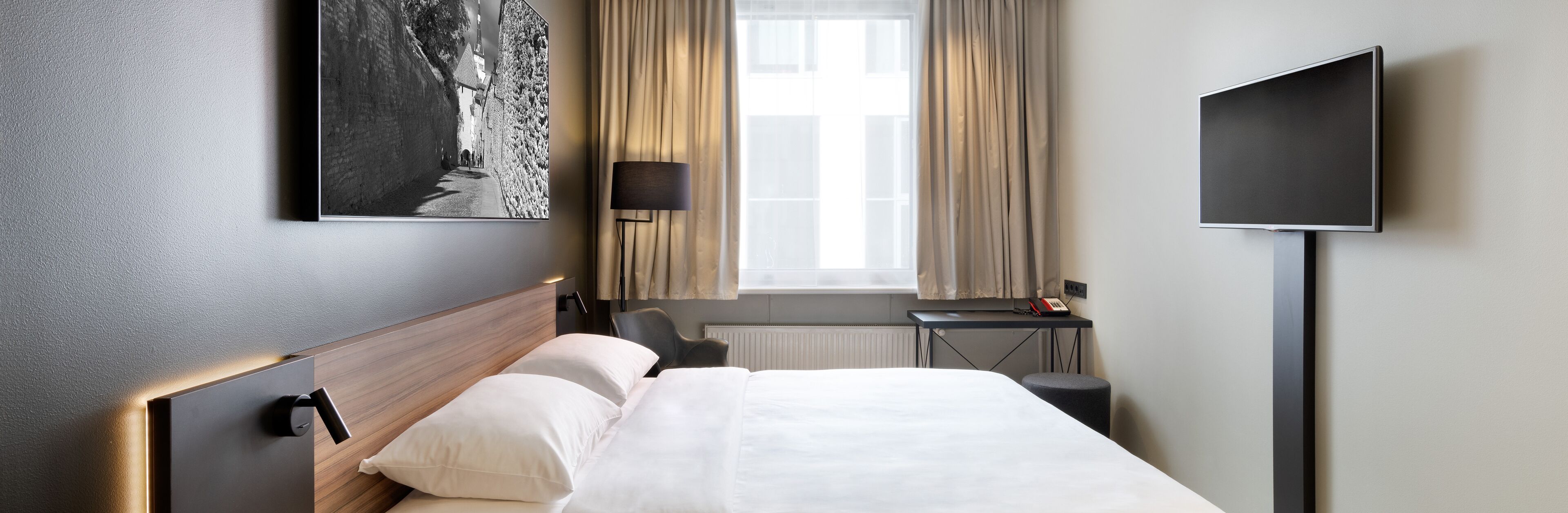 A modern hotel room with a neatly made bed, a desk with a chair, a TV, and a large window with curtains.
