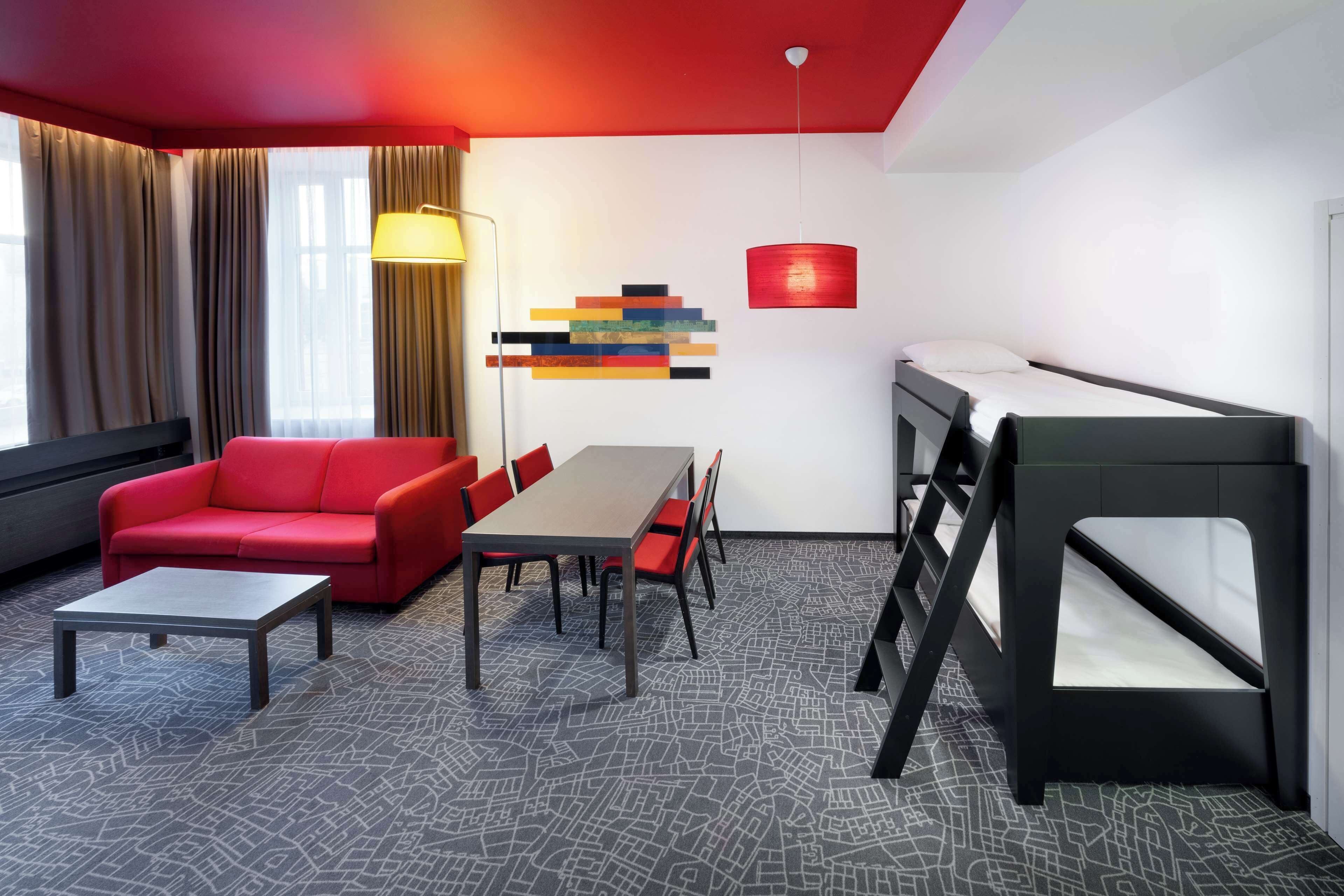 A modern hotel room with a red sofa, a dining table, a bunk bed, and a geometric-patterned carpet.