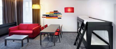 A modern hotel room with a red sofa, a dining table, a bunk bed, and a geometric-patterned carpet.