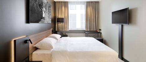 A modern hotel room with a large bed, a flat-screen TV, a desk, and a window with curtains.