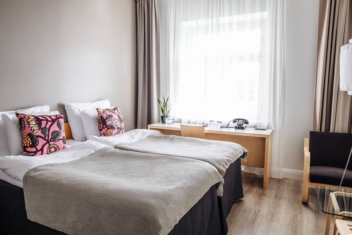 Habitación de hotel con dos camas, un escritorio, una silla y un ventanal con cortinas.