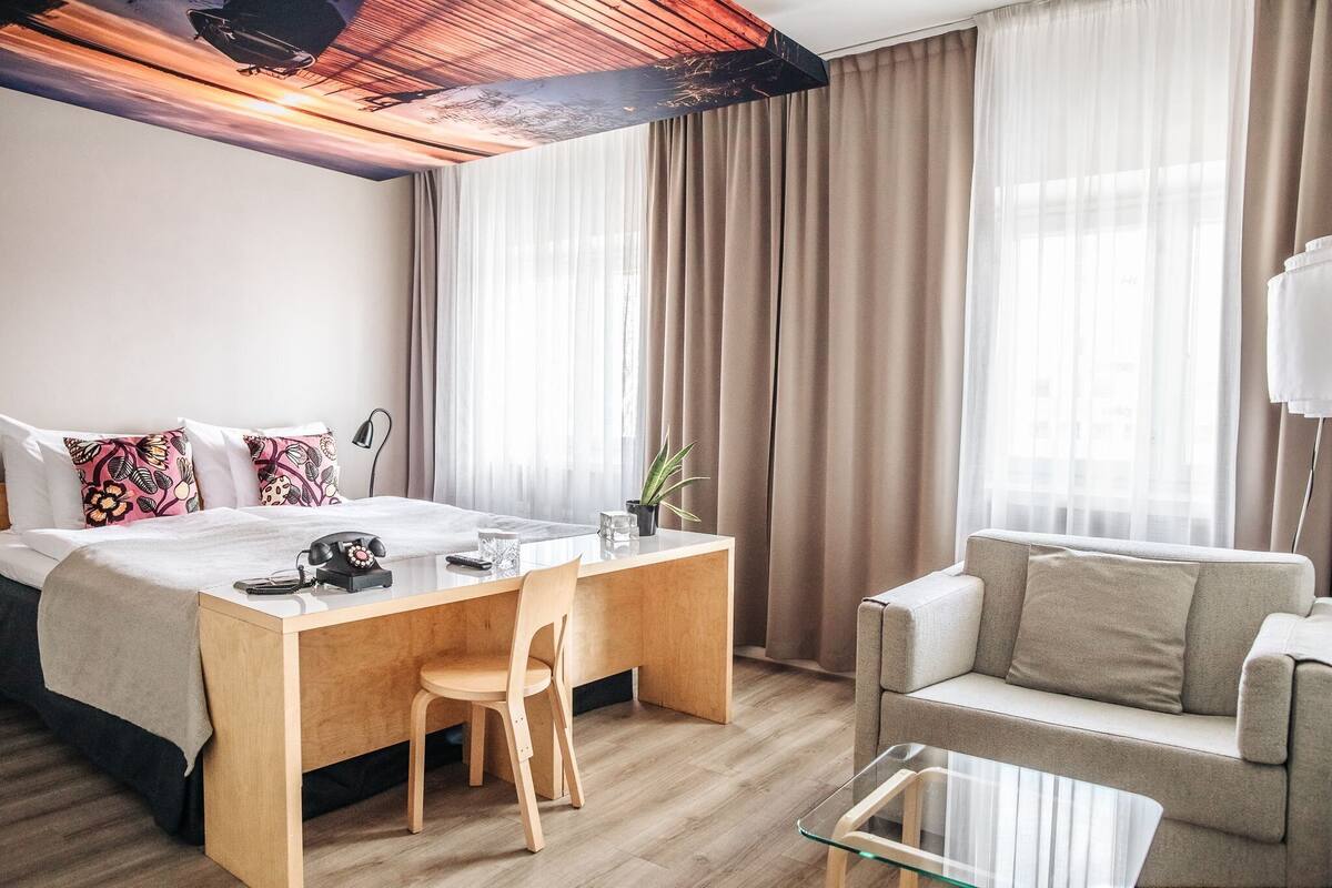 Habitación de hotel con cama, escritorio, silla, sofá y mesa de centro de vidrio.