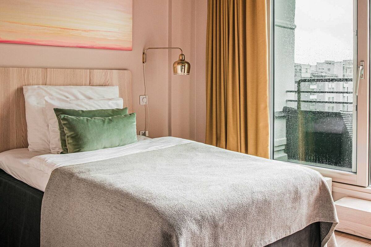 Habitación de hotel con cama, mesita de noche, lámpara, ventana con cortinas y vista a edificios.