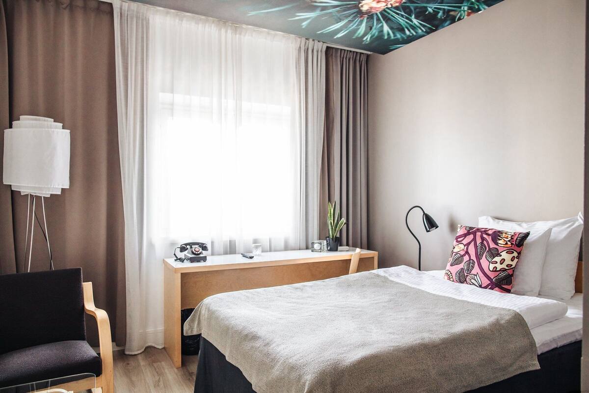 Una habitación de hotel con cama, escritorio, silla, lámpara y ventana con cortinas.