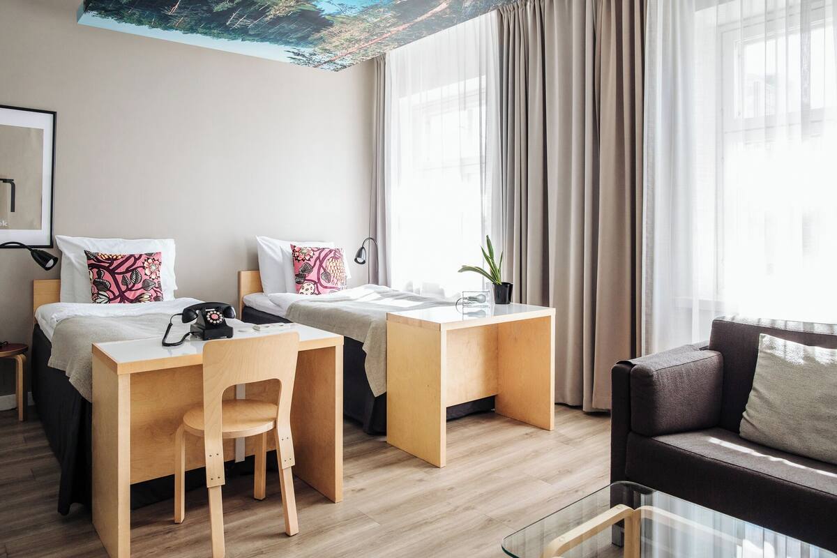 Habitación de hotel con dos camas, un escritorio, una silla, un sofá y un ventanal con cortinas.