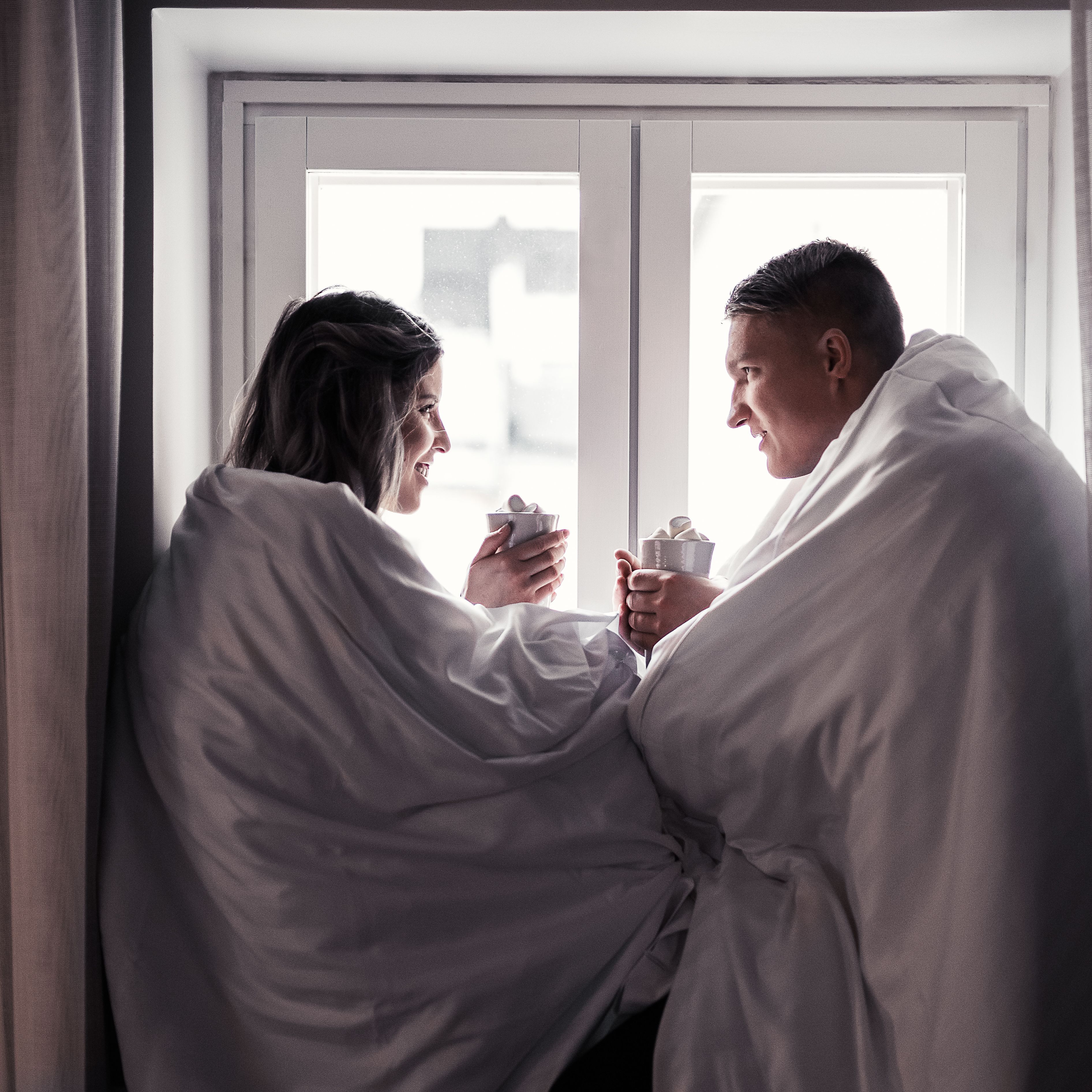 A couple in a cozy room with a large window, holding cups, suggesting a relaxed setting.