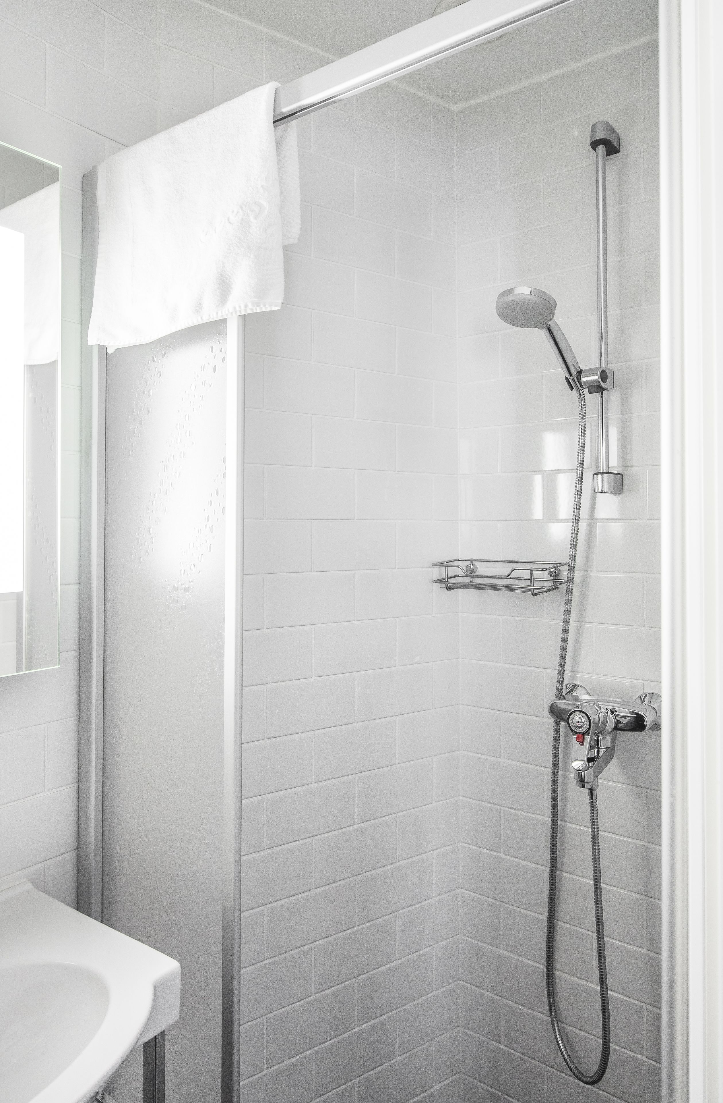 Un baño moderno con azulejos blancos, ducha con cabezal de ducha manual, toallero y un pequeño estante.