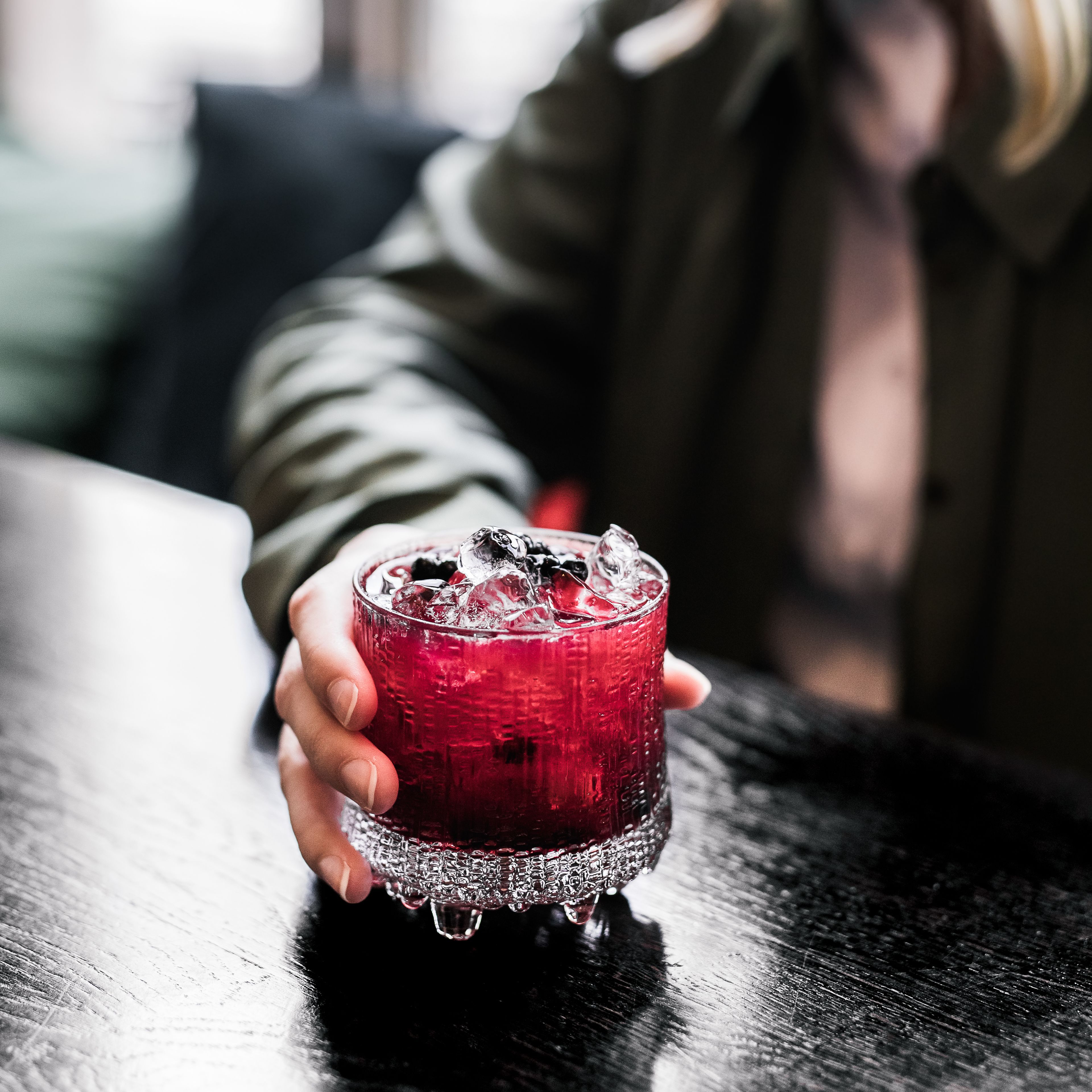 A hand holding a glass with a red beverage, ice cubes, and a garnish, placed on a table.