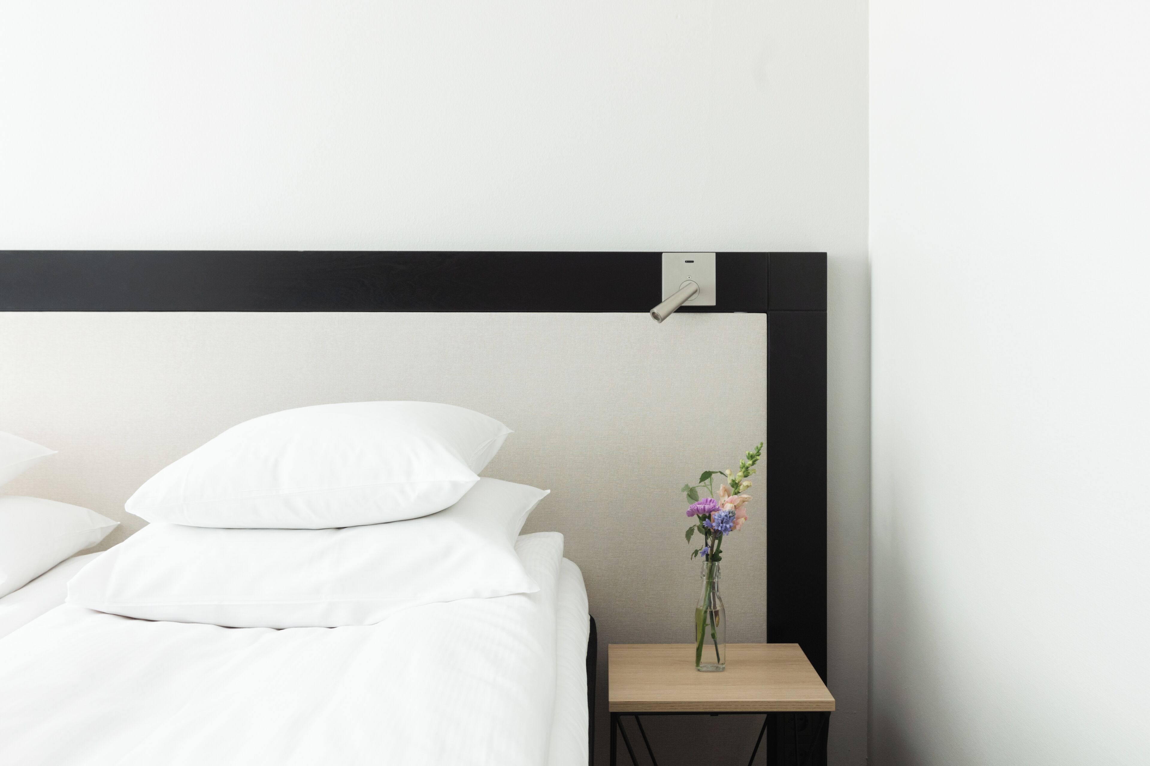 Cama bem arrumada com roupa de cama e almofadas brancas, uma pequena mesa de cabeceira de madeira com um vaso de flores e uma cabeceira preta em frente a uma parede lisa.