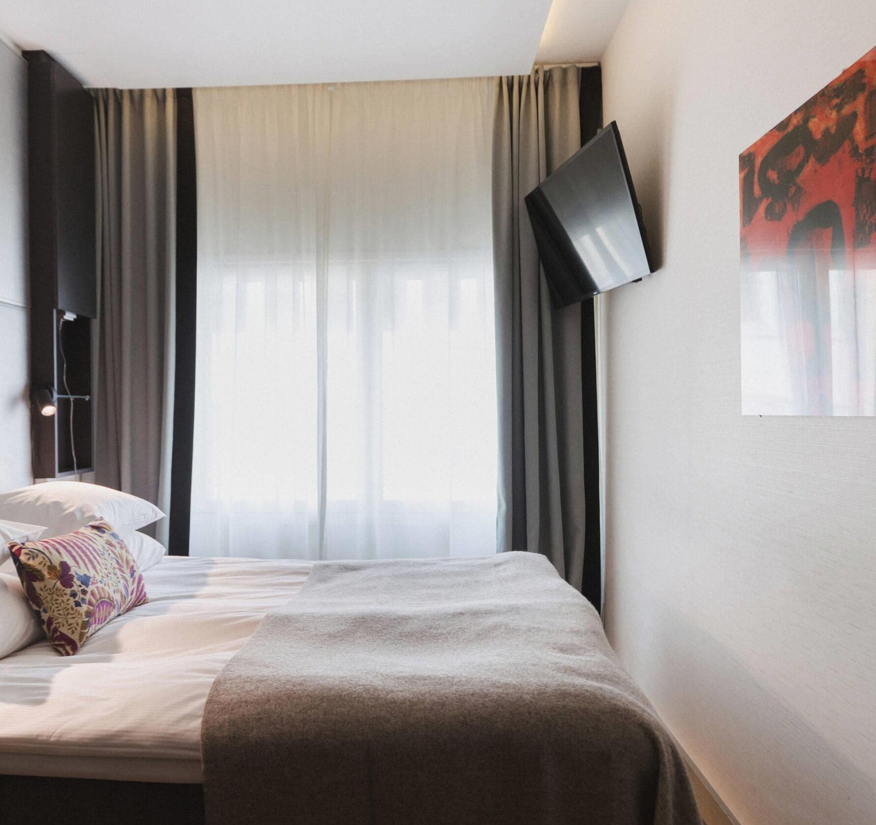 A hotel room with a bed, a flat-screen TV mounted on the wall, and a window with sheer curtains.