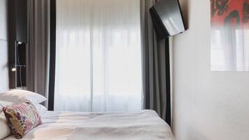 A hotel room with a bed, a flat-screen TV mounted on the wall, and a window with sheer curtains.