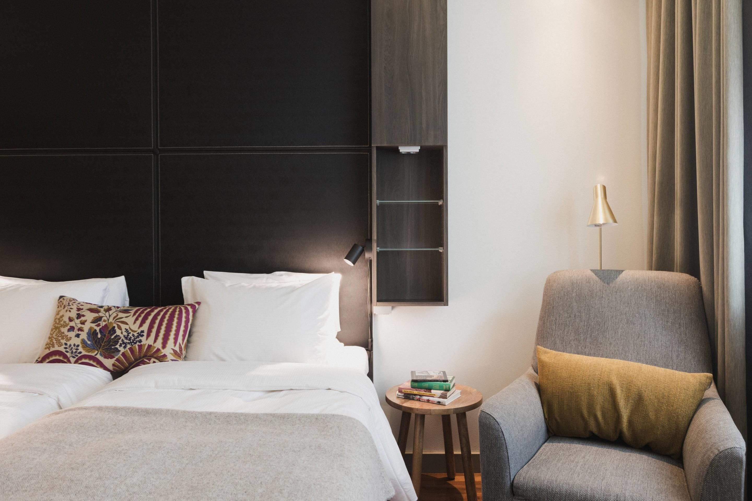 A modern hotel room with a large bed, a small side table, a grey armchair, and a lamp.