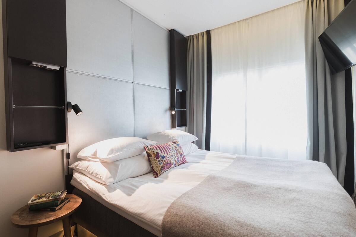 Une chambre d’hôtel moderne avec un lit soigneusement fait, une petite table de chevet sur laquelle repose un livre, et une grande fenêtre avec des rideaux.