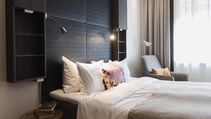 A modern hotel room with a large bed, a bedside table with books, a chair, and a wall-mounted shelf. - Home Hotel Jugend (Helsinki)