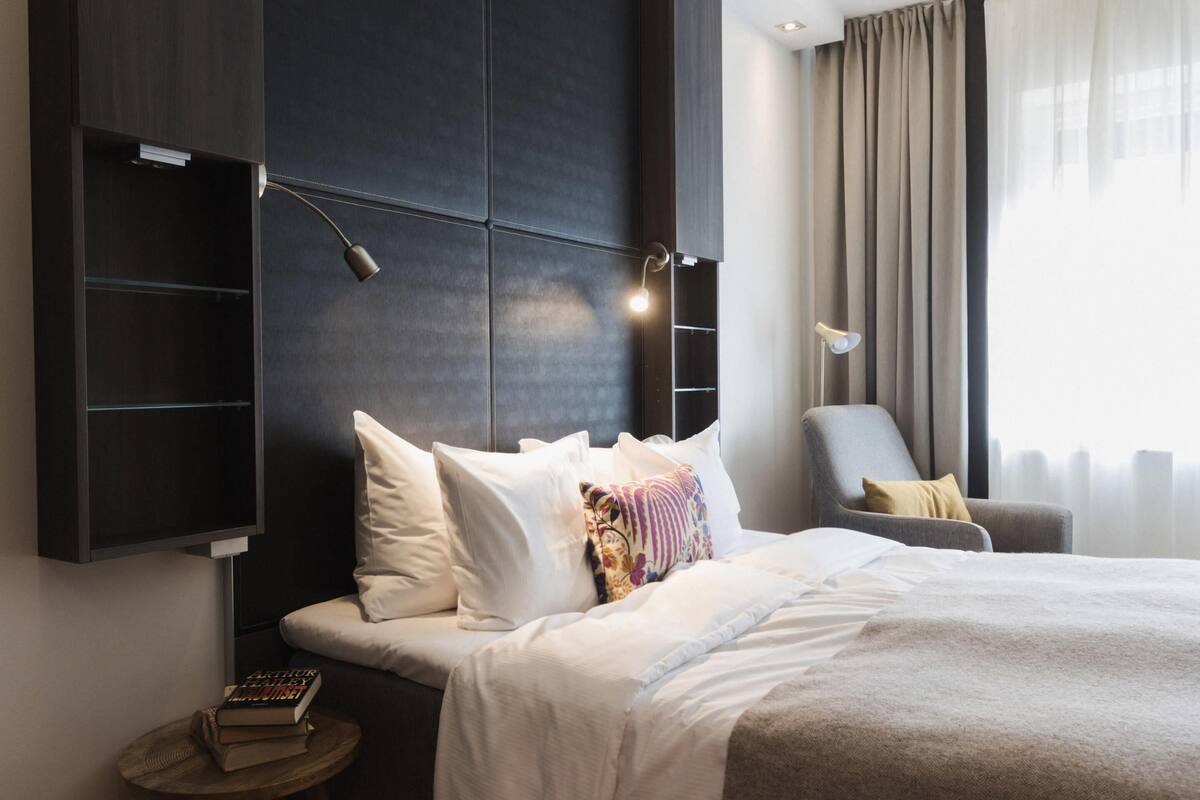 Une chambre d’hôtel moderne avec un grand lit, une table de chevet avec des livres, une chaise et une étagère fixée au mur.