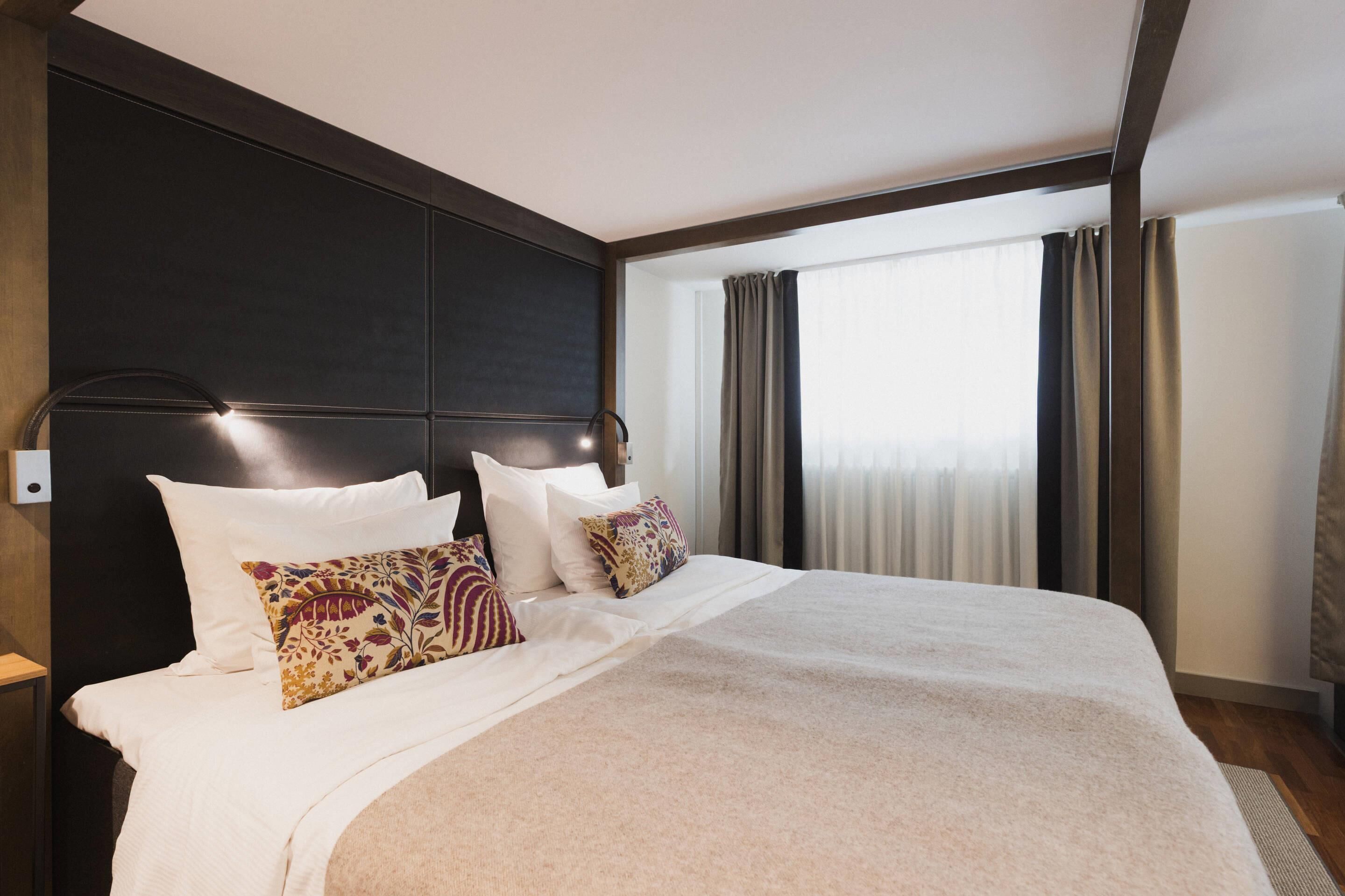 A modern hotel room with a large bed, dark headboard, and a view of the city through large windows.