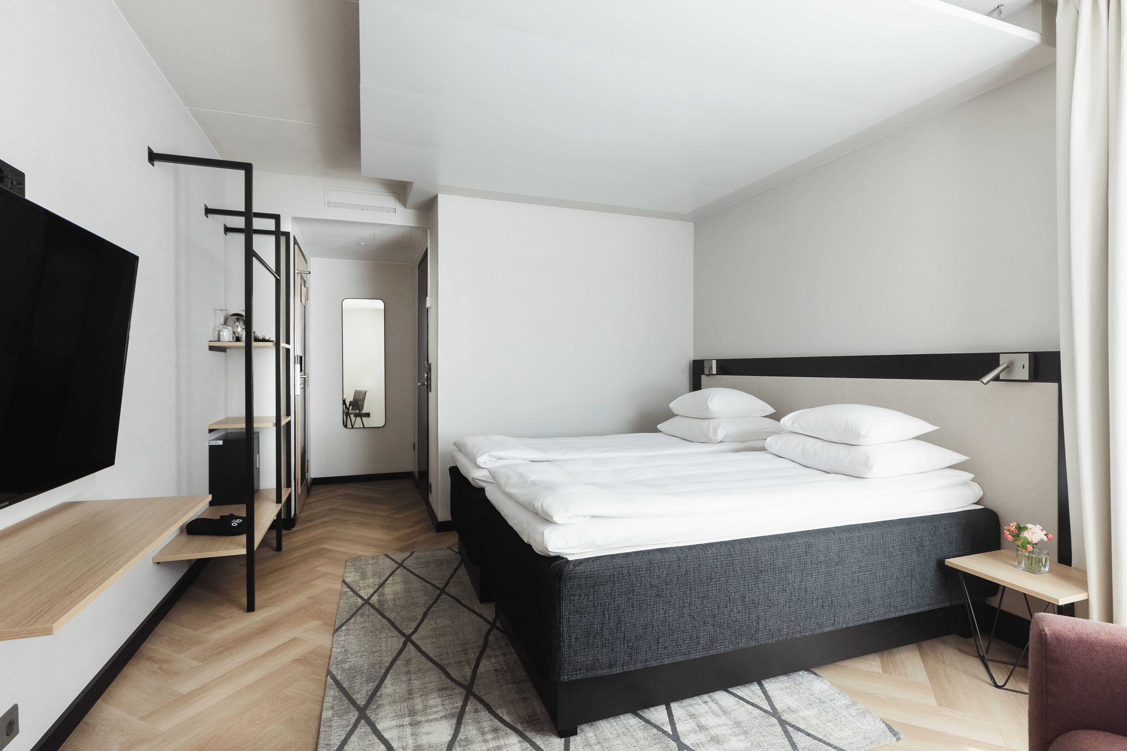 Ett modernt hotellrum med en säng, en TV, ett skrivbord och ett litet bord med en vas.