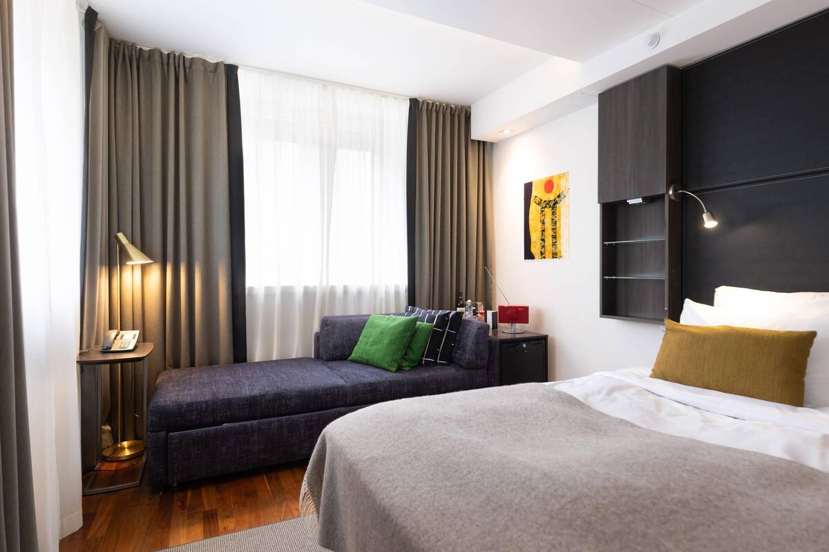 Une chambre d’hôtel avec un lit, un canapé, un bureau avec une lampe et une fenêtre avec des rideaux.