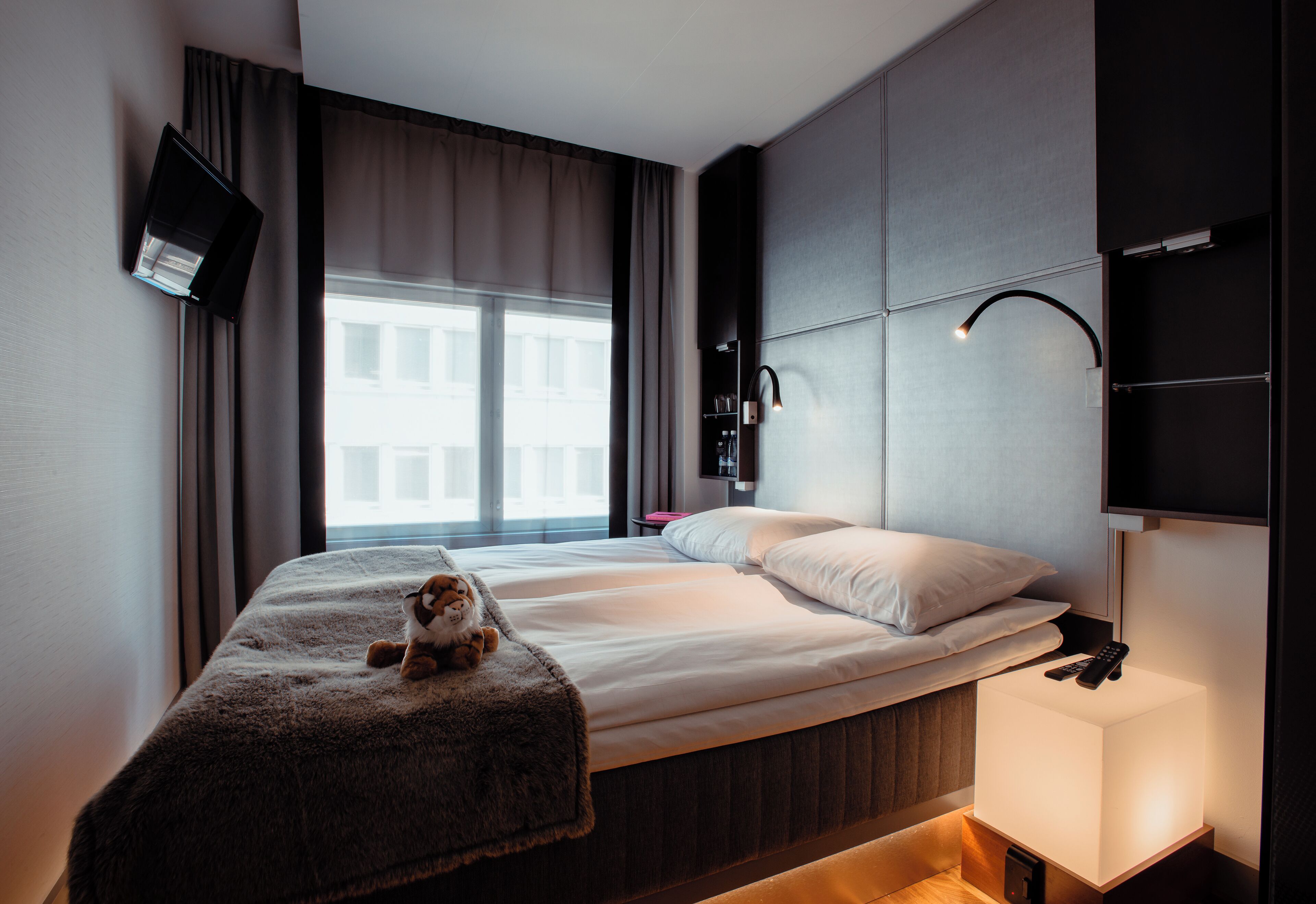 Una habitación de hotel moderna con una cama grande, un televisor montado en la pared, una lámpara de noche y un ventanal con cortinas.